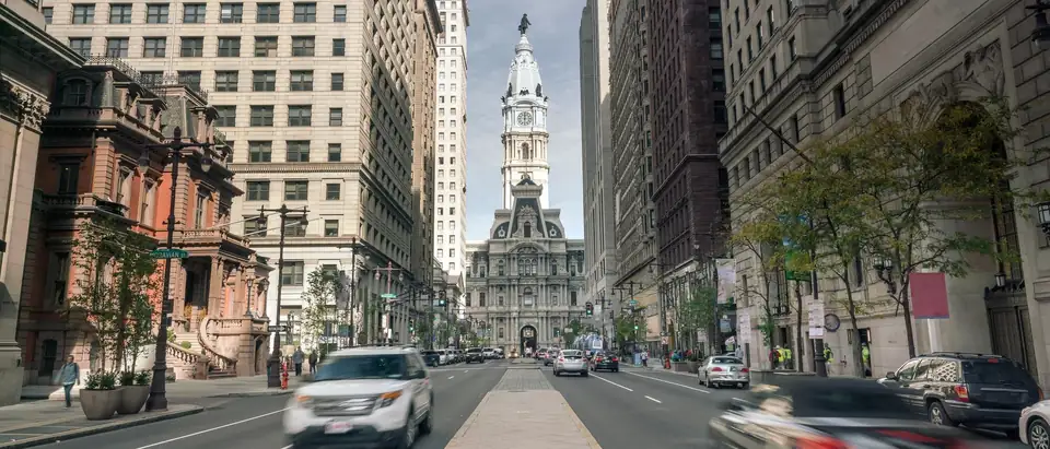 Cars moving along South Broad Street toward Philadelphia City Hall and its clock tower, between tall stone office buildings