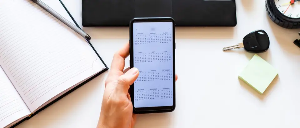 A hand holding a phone that shows a calendar, over a white desk with an open notebook and pen, a car key, a keyboard and a clock