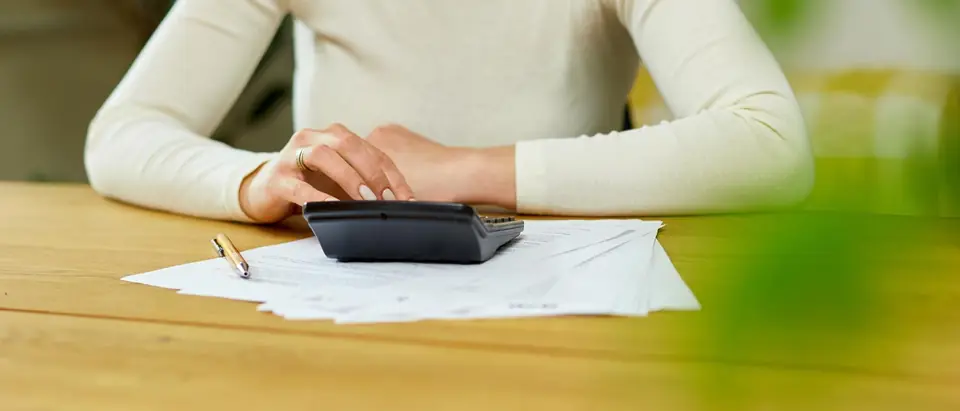 A person seated at a wooden table pressing the keys of a calculator that rests on a spread of printed papers, with a pen beside them