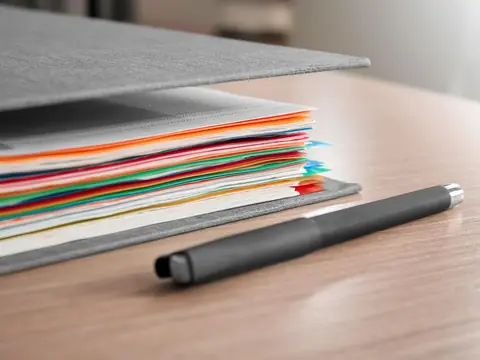 A gray folder holding a thick stack of papers with colored tabs, with a black pen lying beside it on a desk