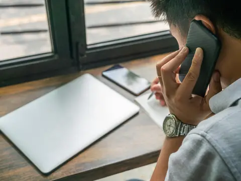 A person seen from behind holding a phone to their ear at a wooden counter by a window, with a closed laptop and a notebook in front of them