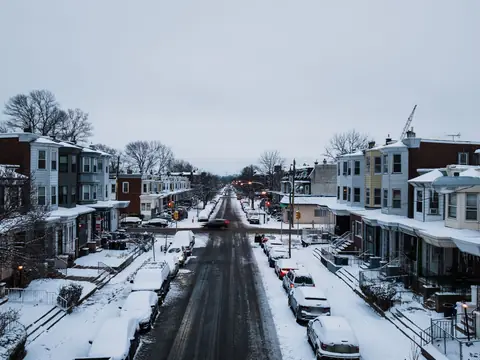 Snow-covered cars parked bumper to bumper along both curbs of a Philadelphia street lined with porch-front rowhouses
